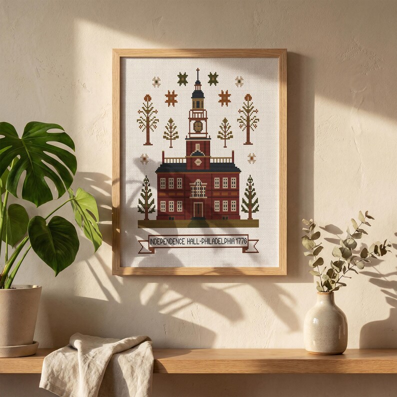 May include: Cross-stitch of Independence Hall in Philadelphia, Pennsylvania, framed in a light wood. The building is rendered in shades of red and brown, with a detailed steeple and surrounding trees. The text "INDEPENDENCE HALL-PHILADELPHIA 1776" is stitched below.
