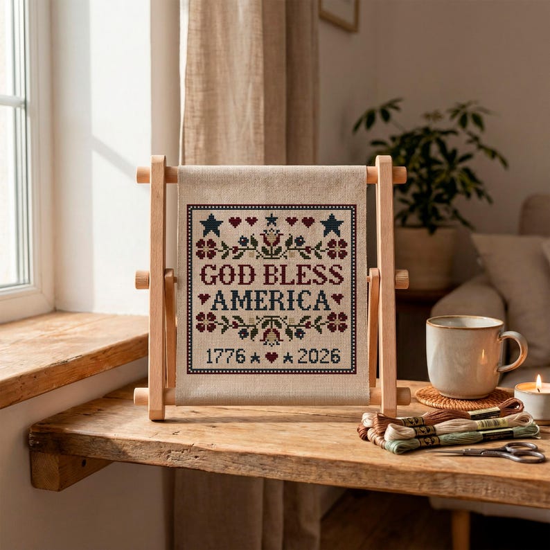 May include: A cross-stitch sampler with the text "GOD BLESS AMERICA" and the dates "1776" and "2026". The sampler is displayed on a wooden frame. The design includes stars, hearts, and floral elements in red, blue, and green.