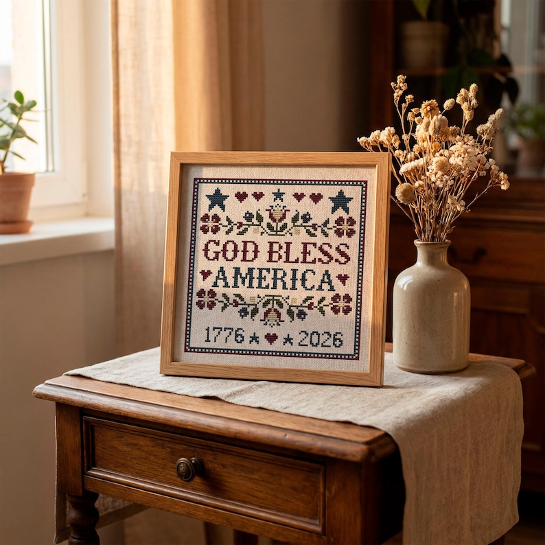 May include: A framed cross-stitch with the words "GOD BLESS AMERICA" in red and black. The design includes stars, hearts, and floral elements. The year "1776" and "2026" are also included. The frame is made of light-colored wood.