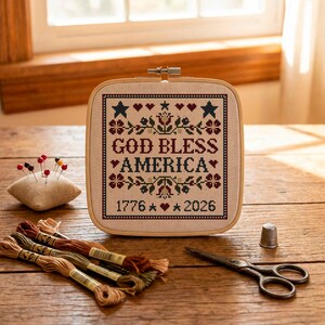 May include: A cross-stitch embroidery hoop with the words "GOD BLESS AMERICA" and the date 1776-2026. The design includes stars, hearts, and floral elements in red, blue, and green. Embroidery floss, a needle cushion, thimble, and scissors are also present.