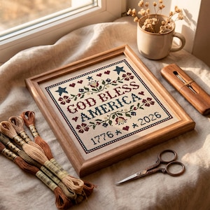 May include: A framed cross-stitch sampler with the text "GOD BLESS AMERICA 1776 * 2026" in red, blue, and green. The wooden frame surrounds the needlework. Embroidery threads and small scissors are also visible.