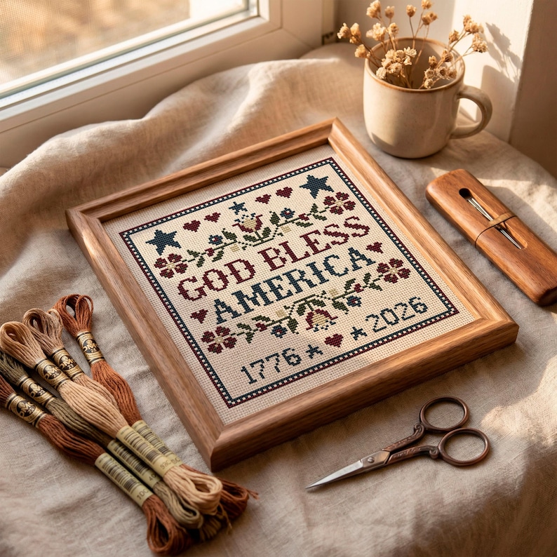 May include: A framed cross-stitch sampler with the text "GOD BLESS AMERICA 1776 * 2026" in red, blue, and green. The wooden frame surrounds the needlework. Embroidery threads and small scissors are also visible.