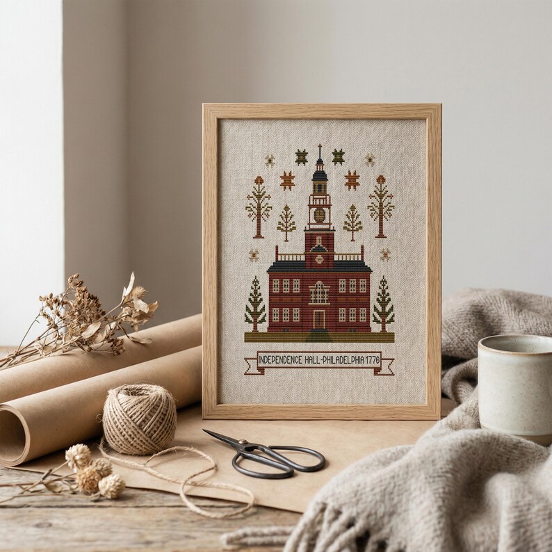 May include: Framed cross-stitch of Independence Hall, Philadelphia, 1776. The building is rendered in shades of red, brown, and green, with a detailed steeple and surrounding trees. The frame is light wood.