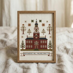 May include: Cross-stitch artwork of Independence Hall in Philadelphia, Pennsylvania, framed in a wooden frame. The building is depicted in shades of red and brown, with a clock tower and surrounding trees. The text "INDEPENDENCE HALL-PHILADELPHIA 1776" is displayed on a banner.