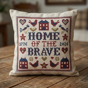 May include: A square, beige pillow with a cross-stitched design. The design features the words "HOME OF THE BRAVE 1776 2026" in red and blue, surrounded by houses, birds, hearts, and stars. The pillow is on a wooden surface.