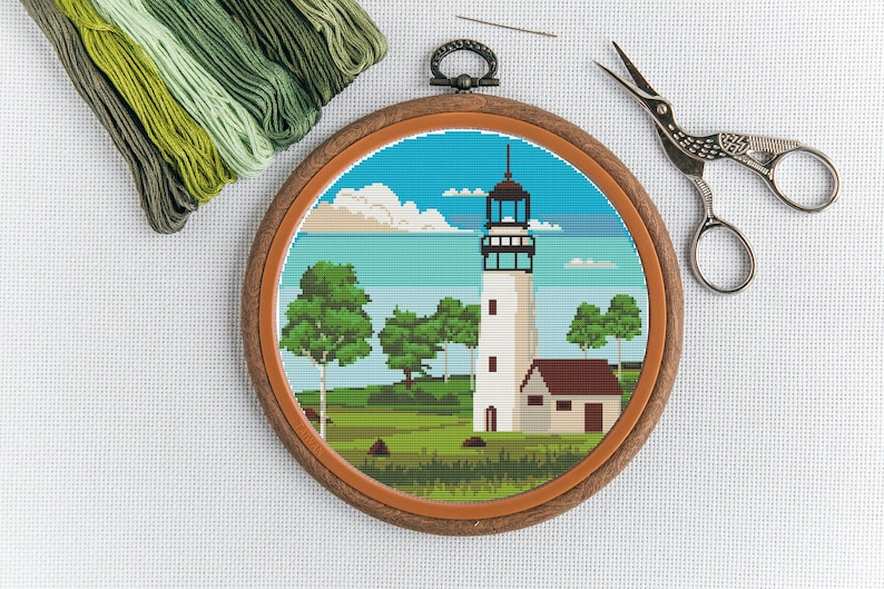 May include: A completed cross-stitch embroidery project depicting a white lighthouse with a brown roof, surrounded by green trees and a blue sea. The embroidery is framed in a brown wooden hoop.