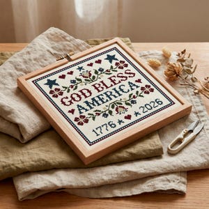 May include: A framed cross-stitch featuring the text "GOD BLESS AMERICA", the dates "1776" and "2026", and decorative stars, hearts, and floral motifs. The design uses red, blue, and green threads. The frame is made of light wood.