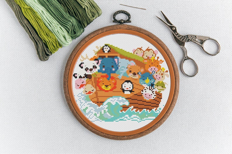 May include: A completed cross-stitch embroidery project in a wooden hoop, depicting a colorful Noah's Ark with animals on board. The ark is surrounded by blue and green waves. The hoop is on a white background with green embroidery floss and a pair of embroidery scissors.