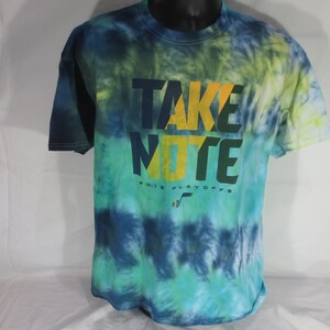 Op de afbeelding: Een tie-dye T-shirt met een groen, blauw en geel ontwerp. Het shirt heeft de tekst "TAKE NOTE" in gele en groene letters met de woorden "SOCCER PLAYOFFS" eronder.