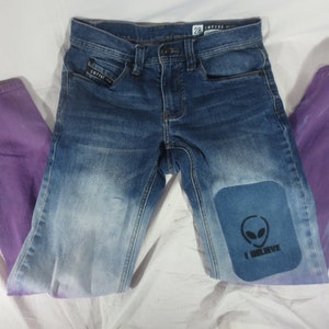 Puede incluir: Un par de pantalones vaqueros azules con un tinte degradado morado en las piernas. La rodilla derecha tiene un parche con un gráfico de extraterrestre y el texto "I Believe".