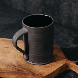 May include: A cylindrical ceramic mug with a matte charcoal gray exterior and a smooth black interior. The mug features a curved handle attached to the side and a slightly flared base, showcasing a simple, functional design for holding beverages.