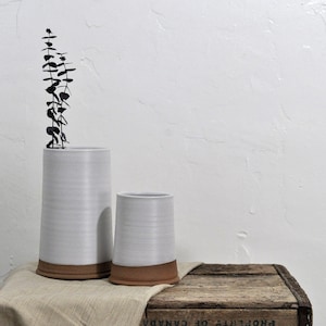 Puede incluir: Dos jarrones de cerámica blanca con bordes marrones, uno más alto que el otro, sobre una superficie de madera con un paño beige. Una rama de eucalipto seco está en el jarrón más alto.