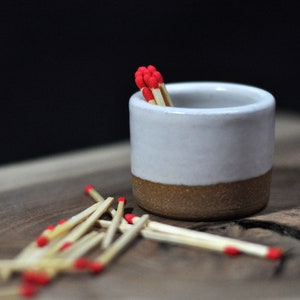 May include: A white ceramic match striker with a brown base, filled with red-tipped matches. The striker is sitting on a wooden surface with scattered matches.