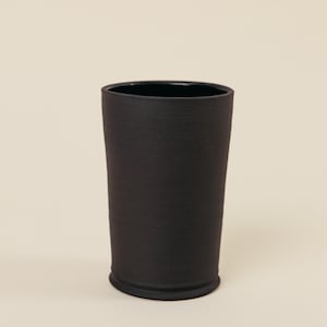 May include: A cylindrical tumbler with a matte black exterior finish and a smooth, dark interior. The vessel features a slightly flared rim and a defined base, presenting a simple, upright form with a consistent dark color throughout the structure.