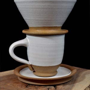 May include: White ceramic coffee dripper with a brown rim, sitting on top of a white ceramic mug with a brown rim and a brown saucer. The mug has a handle. The dripper, mug, and saucer are all on a wooden surface.