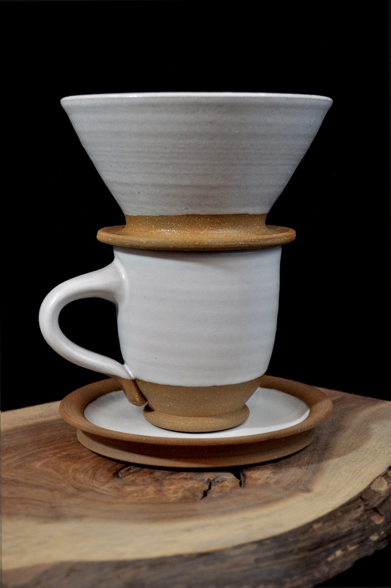 May include: White ceramic coffee dripper with a brown rim, sitting on top of a white ceramic mug with a brown rim and a brown saucer. The mug has a handle. The dripper, mug, and saucer are all on a wooden surface.