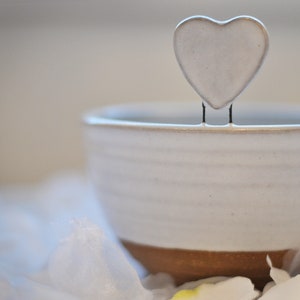 Puede incluir: Un tazón de cerámica blanco con un borde marrón, sentado sobre una superficie de tela blanca. Un objeto blanco en forma de corazón está encaramado en el borde del tazón.