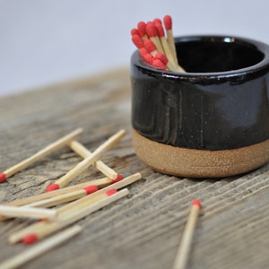 Puede incluir: Un porta cerillas de cerámica negra con un borde marrón, lleno de cerillas con puntas rojas. Cerillas dispersas con puntas rojas están sobre una superficie de madera.