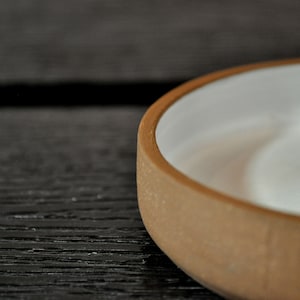 May include: A ceramic ring holder with a white glazed interior and a brown exterior. The holder is shaped like a small bowl with a raised center.