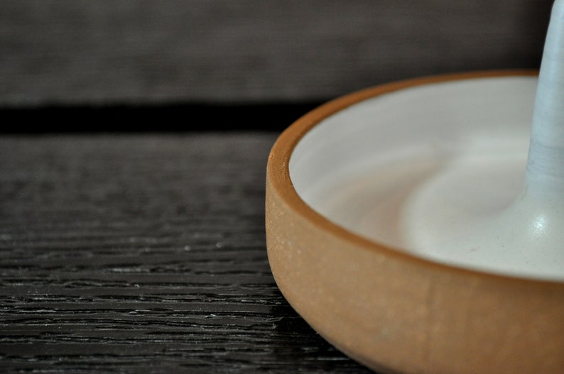 May include: A ceramic ring holder with a white glazed interior and a brown exterior. The holder is shaped like a small bowl with a raised center.