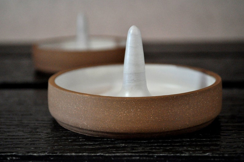 May include: A brown ceramic ring holder with a white ceramic center post. The holder has a speckled brown glaze.