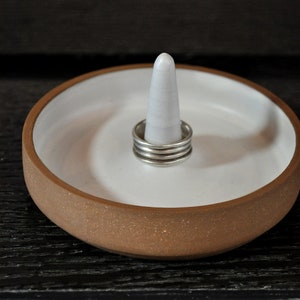 May include: A white ceramic ring dish with a brown rim and a white ceramic cone in the center. The dish has a speckled brown glaze. There are four silver rings stacked on the cone.