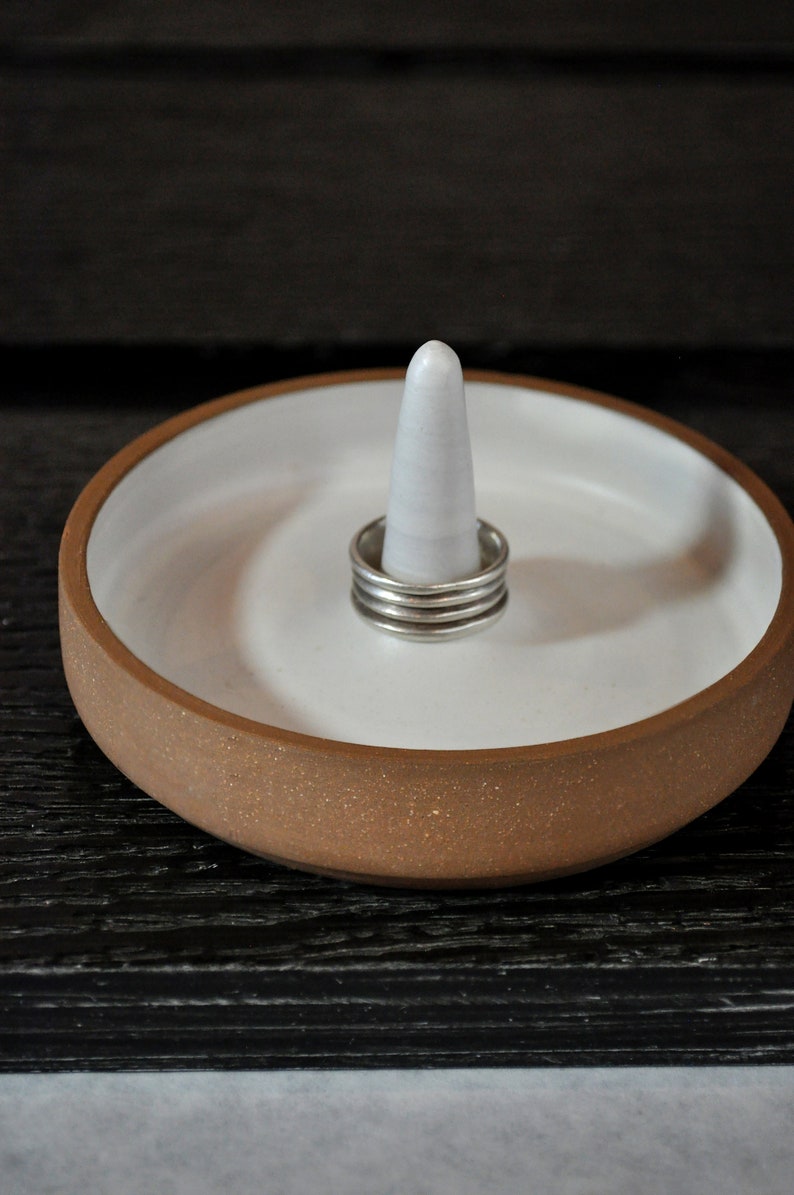 May include: A white ceramic ring dish with a brown rim and a white ceramic cone in the center. The dish has a speckled brown glaze. There are four silver rings stacked on the cone.