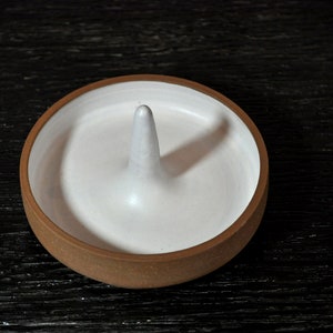 May include: A white ceramic ring holder with a raised center, resting on a dark wooden surface.