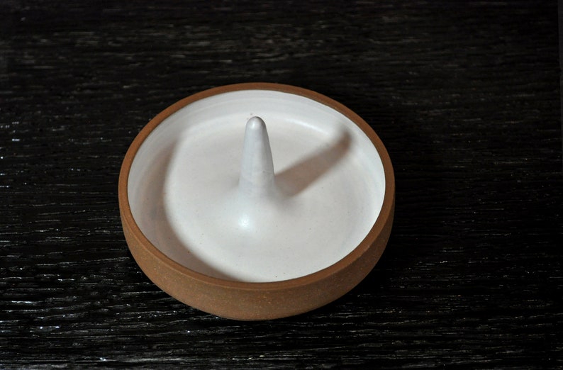 May include: A white ceramic ring holder with a raised center, resting on a dark wooden surface.