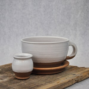 Puede incluir: Una taza de cer&aacute;mica blanca con un borde marr&oacute;n y un platillo a juego, y una taza de cer&aacute;mica blanca m&aacute;s peque&ntilde;a con un borde marr&oacute;n, todo sobre una pieza de madera desgastada.