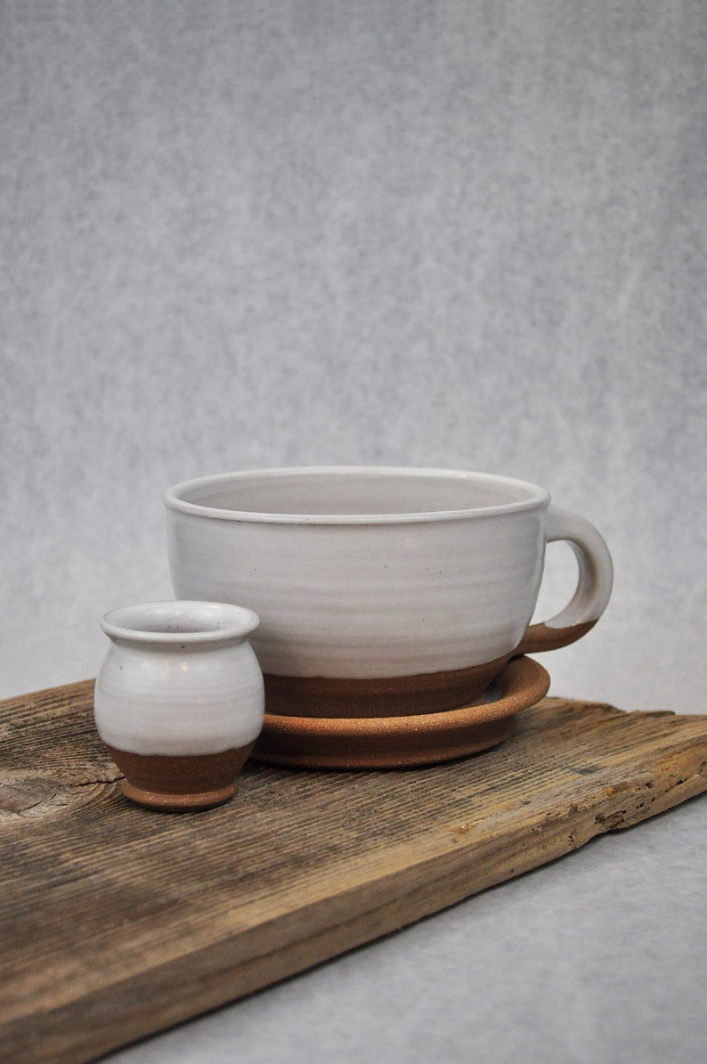 Puede incluir: Una taza de cer&aacute;mica blanca con un borde marr&oacute;n y un platillo a juego, y una taza de cer&aacute;mica blanca m&aacute;s peque&ntilde;a con un borde marr&oacute;n, todo sobre una pieza de madera desgastada.