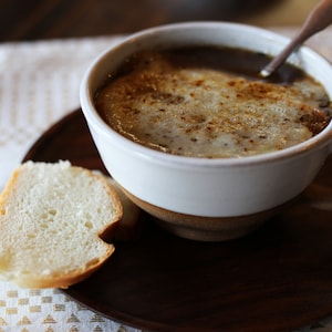 Puede incluir: Un taz&oacute;n blanco de sopa de cebolla francesa con queso derretido encima, servido con una rebanada de pan en una bandeja de madera.