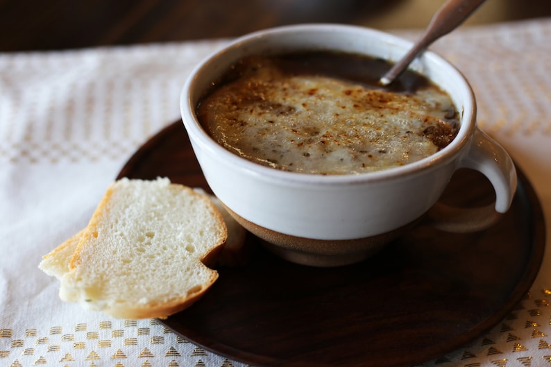 Puede incluir: Un taz&oacute;n blanco de sopa de cebolla francesa con queso derretido encima, servido con una rebanada de pan en una bandeja de madera.