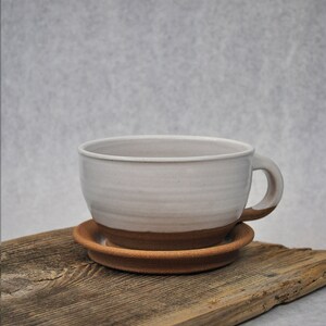 Puede incluir: Una taza de cer&aacute;mica blanca con un borde marr&oacute;n y un platillo a juego. La taza y el platillo est&aacute;n sobre una pieza de madera desgastada.