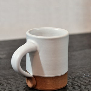 May include: A white ceramic mug with a brown rim and base. The mug has a rounded handle.