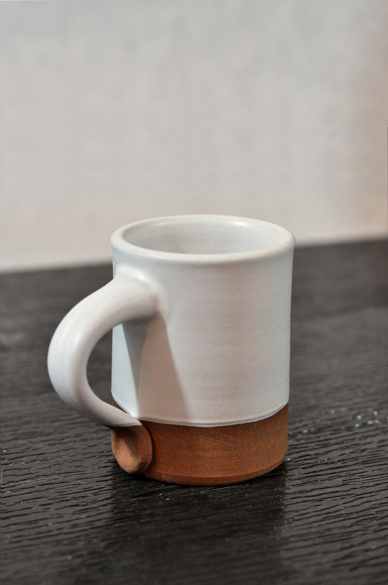 May include: A white ceramic mug with a brown rim and base. The mug has a rounded handle.