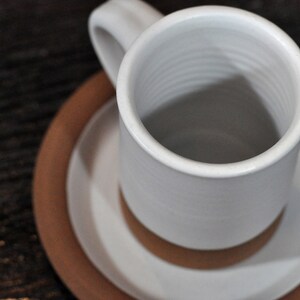 May include: A white ceramic mug with a brown saucer. The mug has a simple design and is empty.