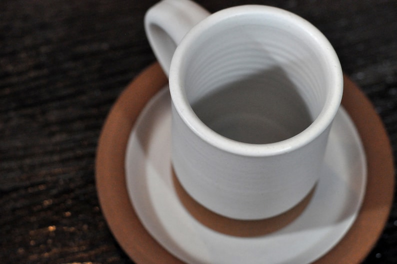 May include: A white ceramic mug with a brown saucer. The mug has a simple design and is empty.