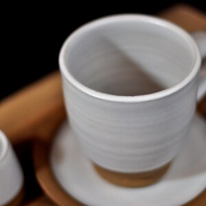 May include: A white ceramic coffee cup with a handle and a white saucer. The cup is sitting on a wooden surface.