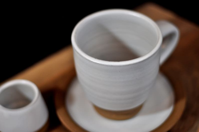 May include: A white ceramic coffee cup with a handle and a white saucer. The cup is sitting on a wooden surface.