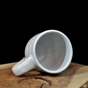 May include: A white ceramic mug with a slightly textured surface and a rounded handle. The mug is sitting on a wooden surface.