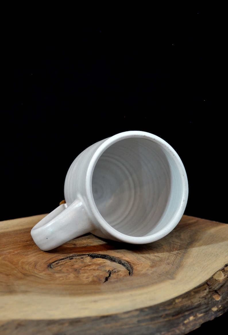 May include: A white ceramic mug with a slightly textured surface and a rounded handle. The mug is sitting on a wooden surface.