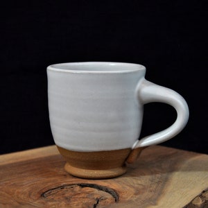 May include: A white ceramic mug with a brown rim and a handle. The mug is sitting on a wooden surface.