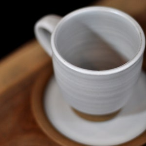 May include: A white ceramic mug with a rounded handle sits on a white saucer. The mug and saucer are on a wooden surface.