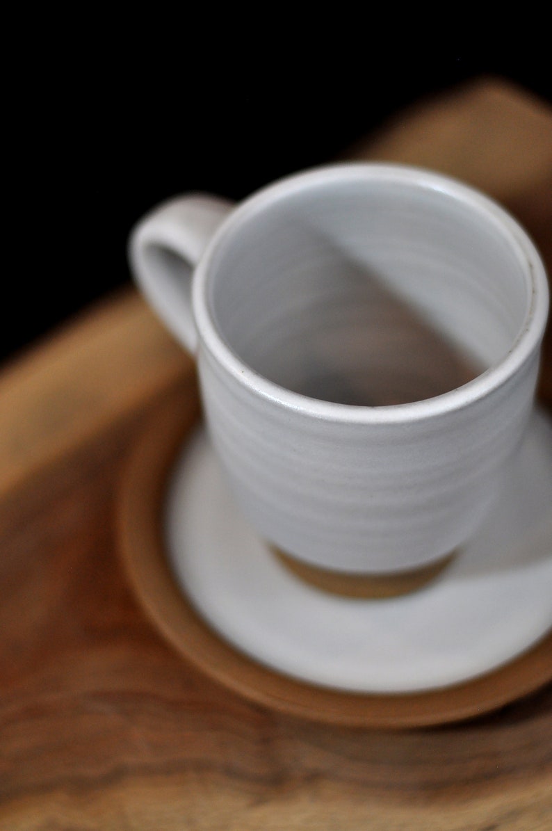 May include: A white ceramic mug with a rounded handle sits on a white saucer. The mug and saucer are on a wooden surface.