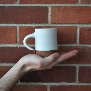 May include: A white ceramic mug with a brown rim, held in a hand against a brick wall background.