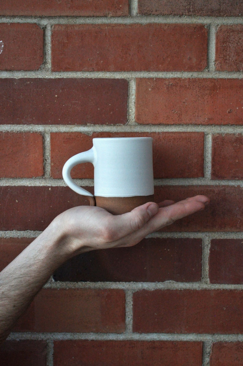 May include: A white ceramic mug with a brown rim, held in a hand against a brick wall background.