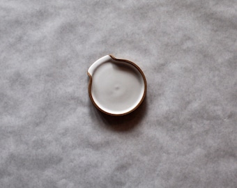 May include: A white ceramic dish with a brown rim. The dish has a unique shape with a small indentation on one side.