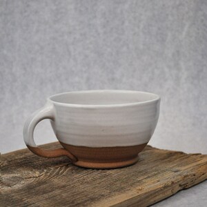 Puede incluir: Una taza de cer&aacute;mica blanca con un borde y un asa marrones. La taza est&aacute; sentada sobre un trozo de madera desgastada.