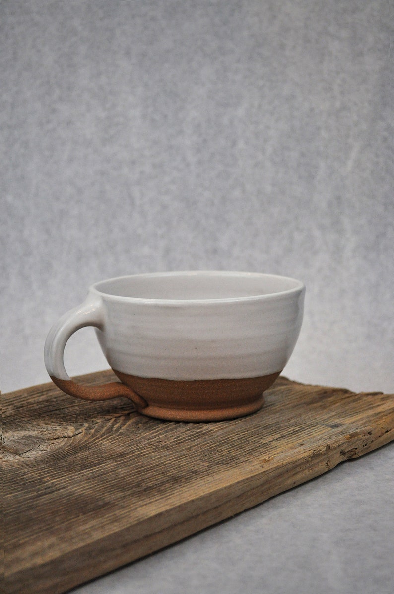 Puede incluir: Una taza de cer&aacute;mica blanca con un borde y un asa marrones. La taza est&aacute; sentada sobre un trozo de madera desgastada.